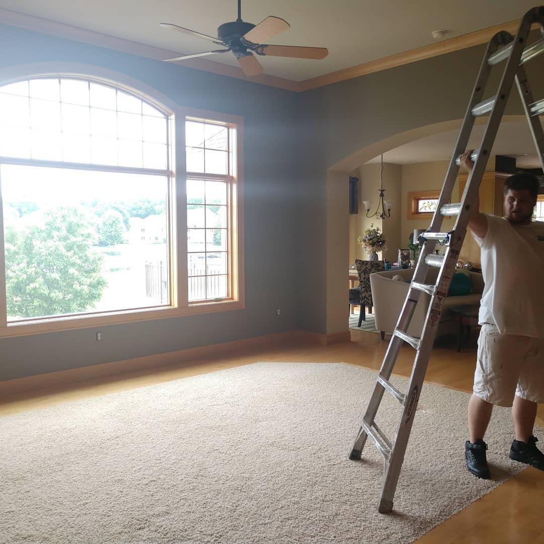 Man holding a ladder inside a room with large windows, light carpet, and olive green walls.