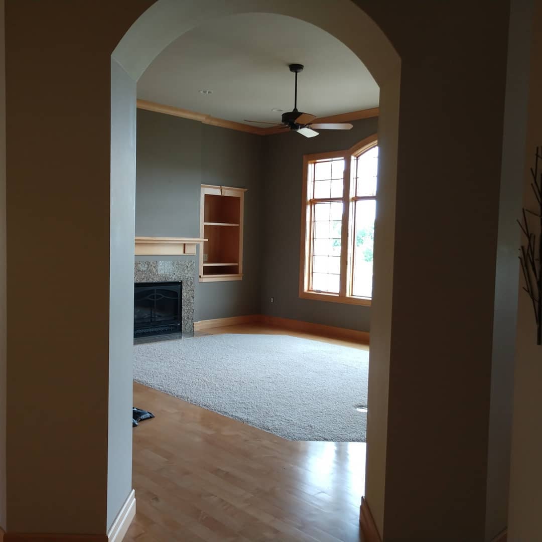Archway into a room with fireplace, built-in shelving, and large windows. Room has carpet and dark walls.
