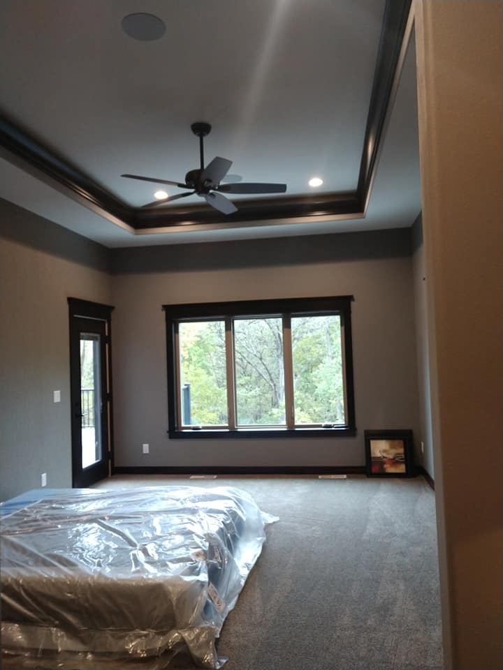 Bedroom with gray walls, a dark ceiling fan, windows, and bed covered in plastic.