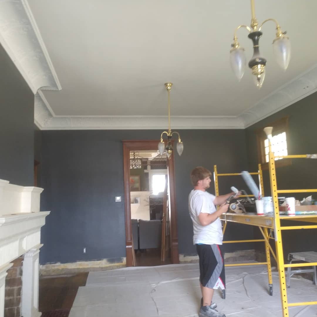 Historic Restoration. Person painting a room with dark gray walls, a white ceiling, and a fireplace.