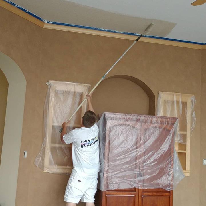 Person paints a ceiling with a roller, standing in a room with a brown wall.