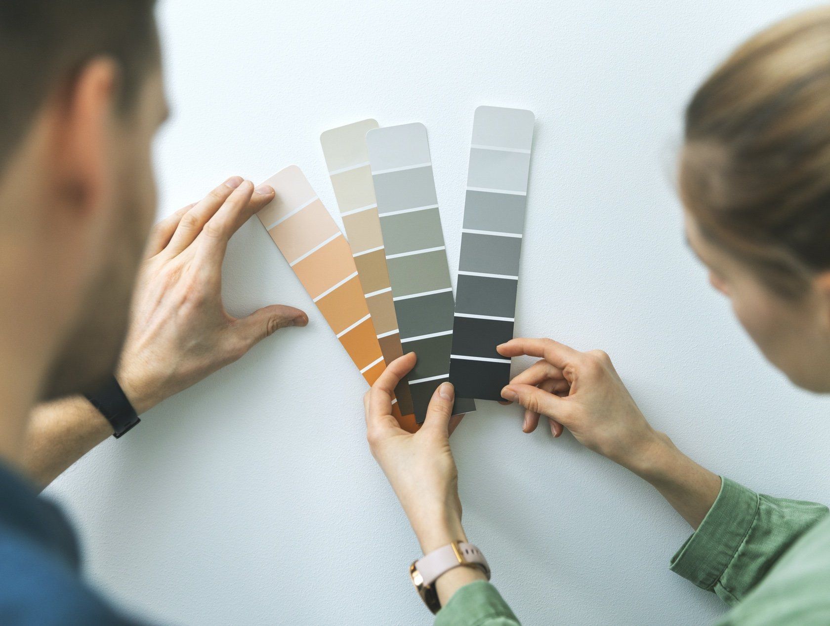 Two people comparing paint swatches on a white wall, choosing a color for interior decorating.