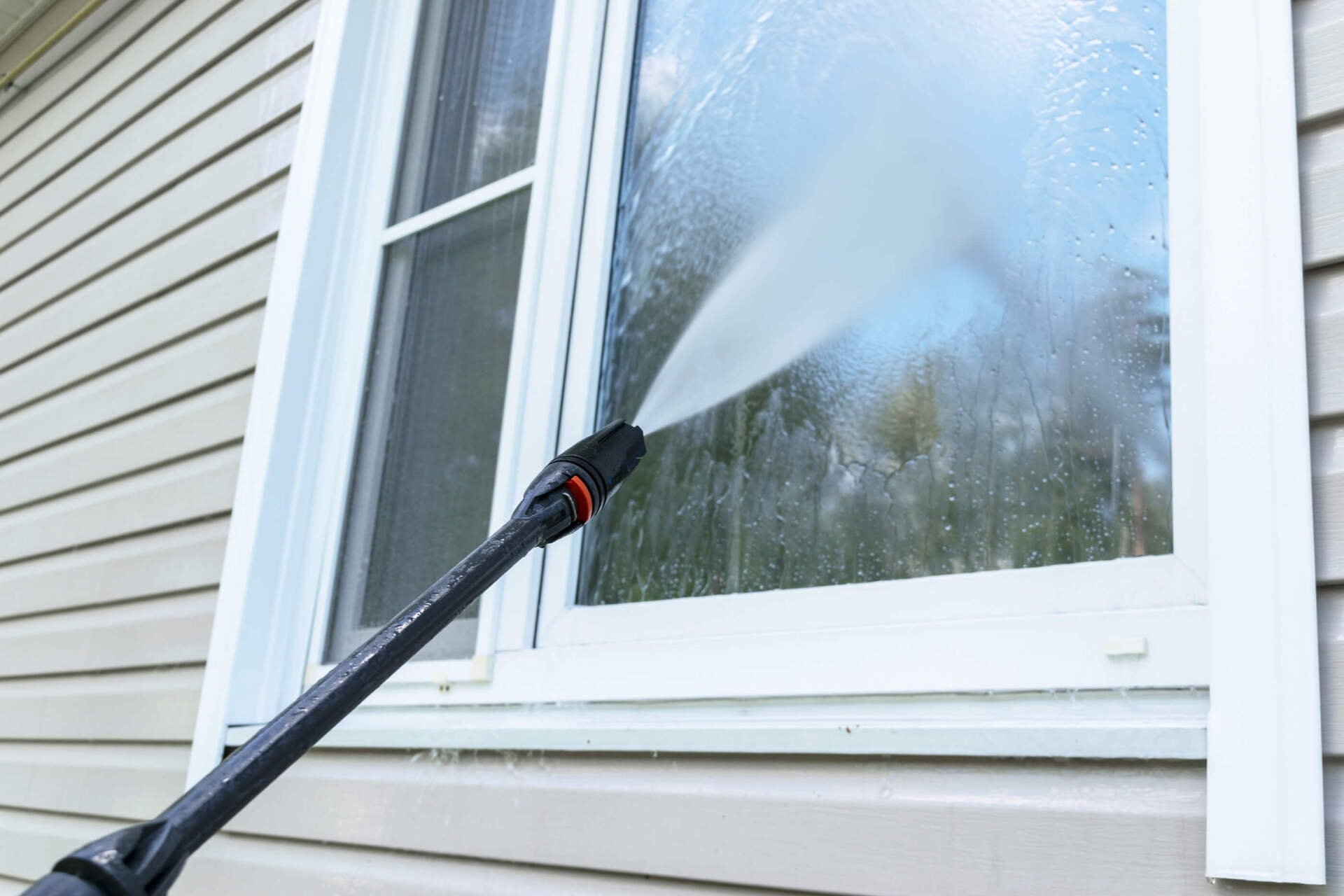Pressure washer spraying a window on a house with beige siding.