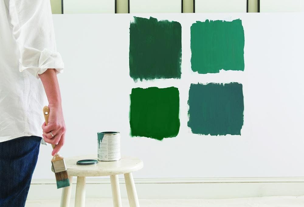 Person holds a paintbrush, evaluating four green paint swatches on a white wall; paint can and stool nearby.