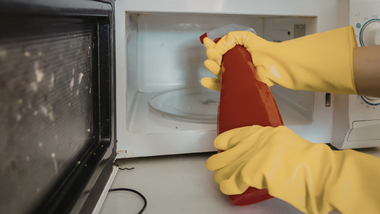 Person wearing yellow gloves sprays cleaning solution inside a microwave.