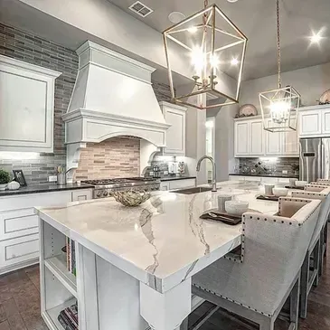 Elegant white kitchen with marble island, gold chandeliers, brick backsplash, and bar seating.