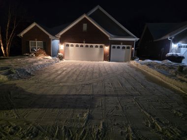 Snow-covered suburban driveway leading to a two-car garage at night, lit by warm porch lights