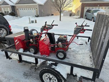 Red and black snow blowers on a trailer in a snowy driveway.