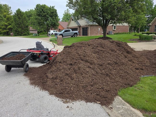 Large mulch pile beside a gravel driveway with a red utility cart and suburban houses in the background