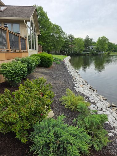 House beside a calm lake with a rocky shoreline and green shrubs under an overcast sky