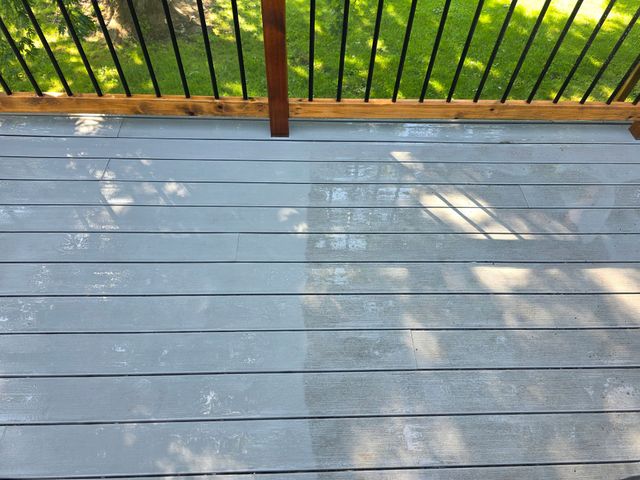 Gray wooden deck with sunlit shadows and a black railing overlooking green trees and grass.