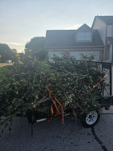 Trailer loaded with cut branches and brush, secured with orange straps on a driveway at dusk