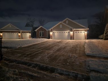 Snowy suburban house at night with lit windows, garage doors, and a dark driveway
