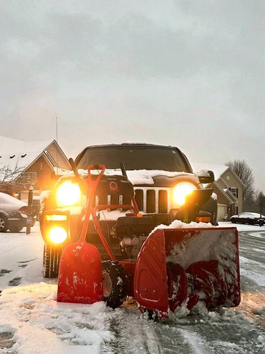 Snowplow clearing snow on a driveway, with bright headlights and red plow blade at dusk