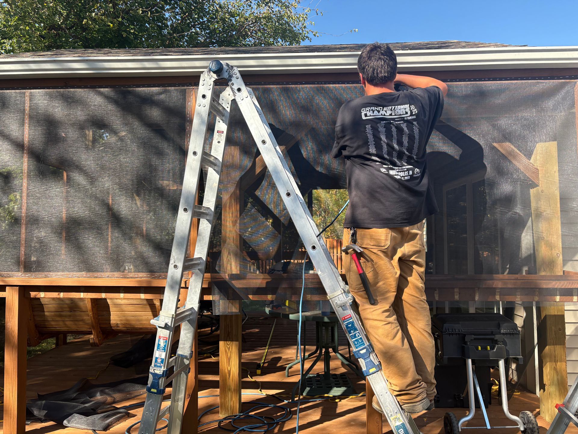 Person on a ladder repairing a screen-covered porch under a roofline.