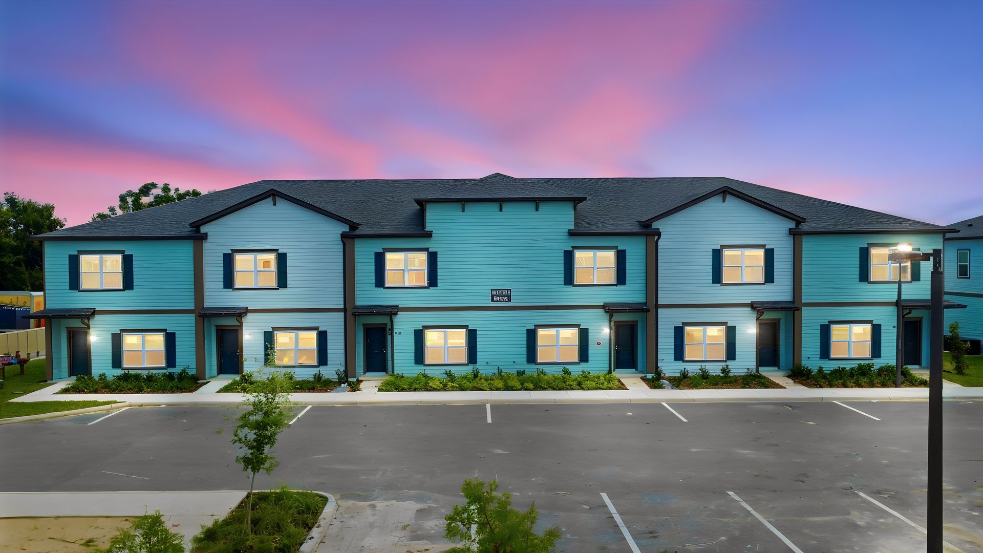 Blue two-story apartment building at dusk with lit windows and empty parking lot under a pink sky