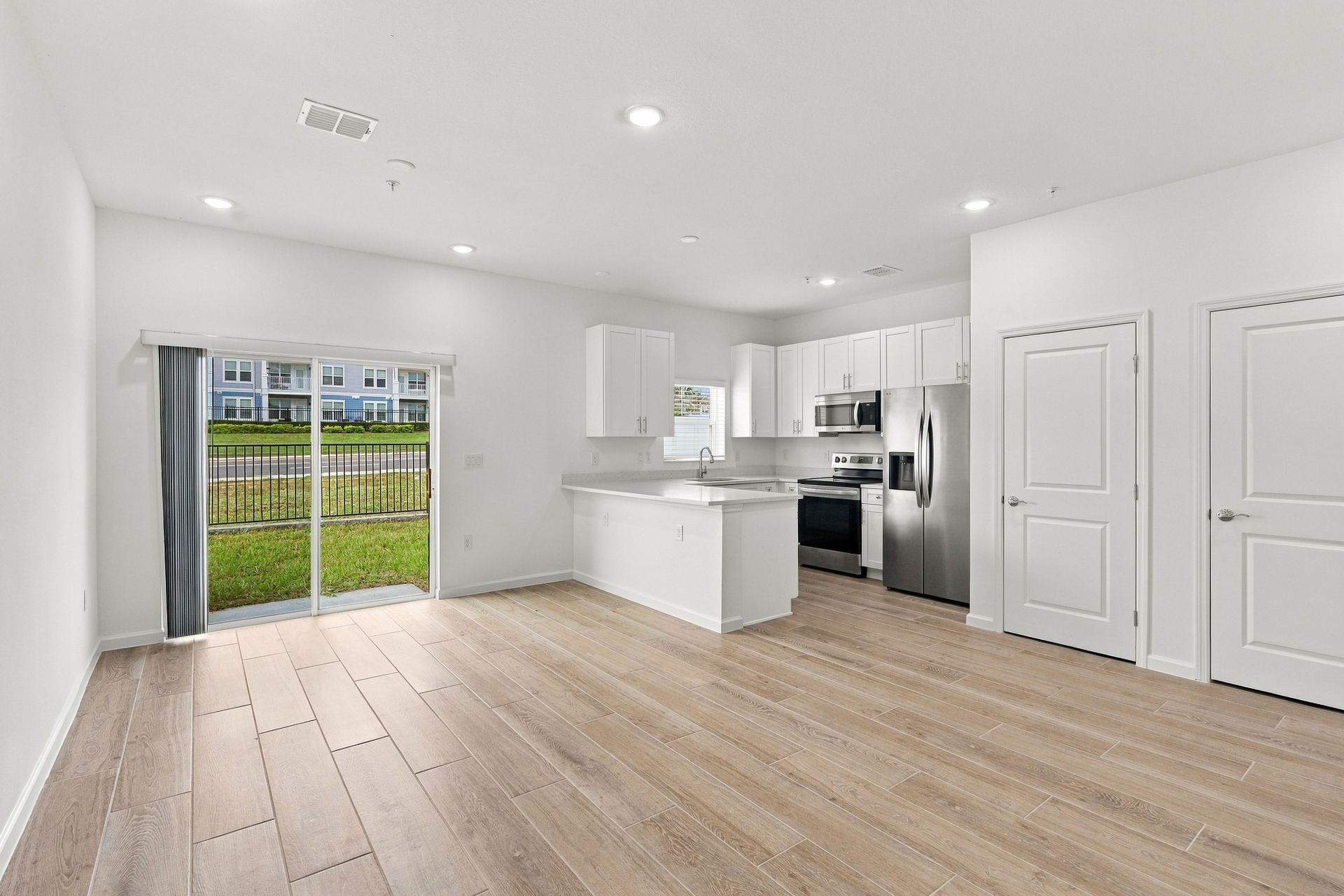 Bright open-concept living room with kitchen, white walls, light wood floors, and sliding glass doors to a yard