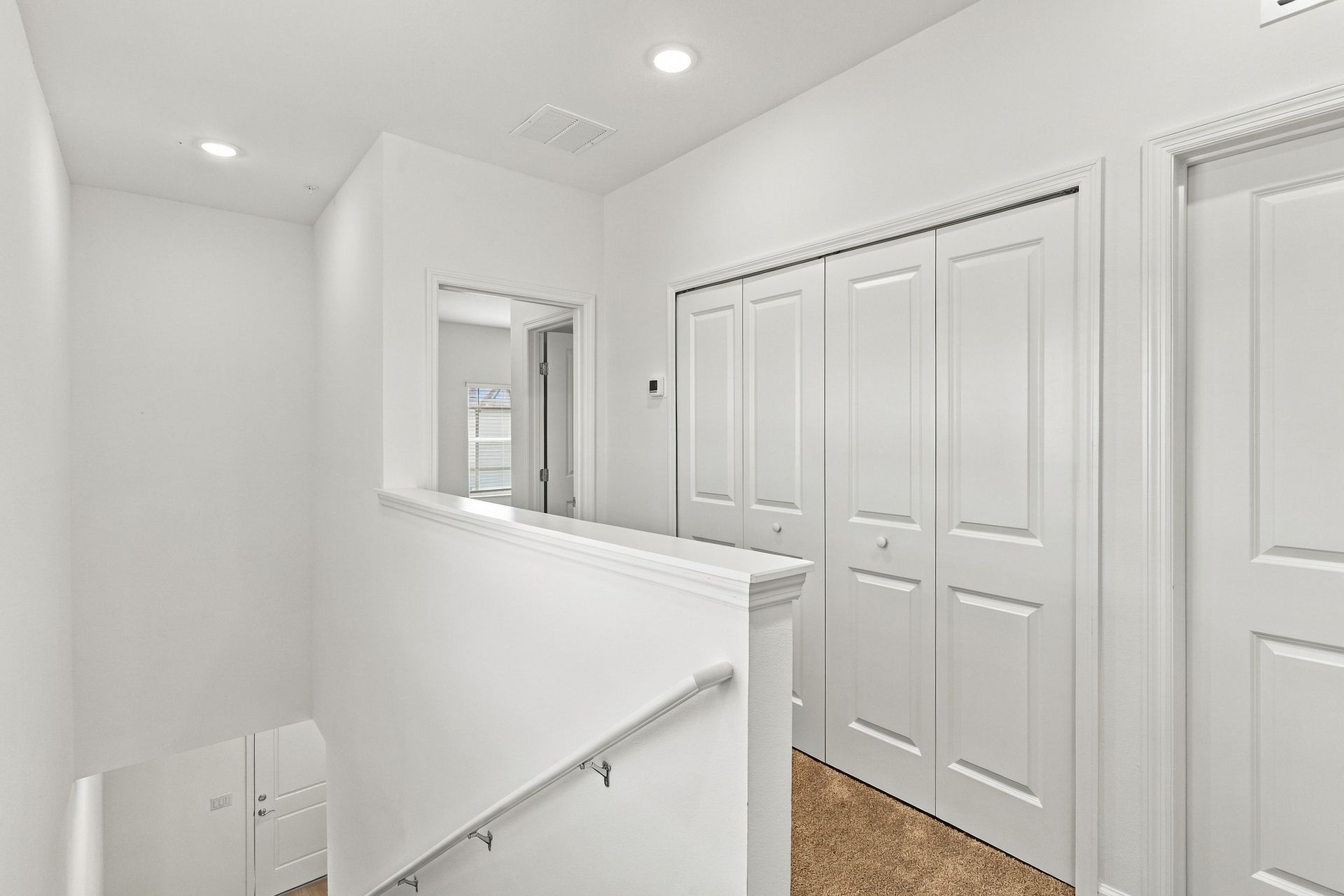 Bright white hallway with closet doors, a stair railing, and carpeted floor under recessed lights