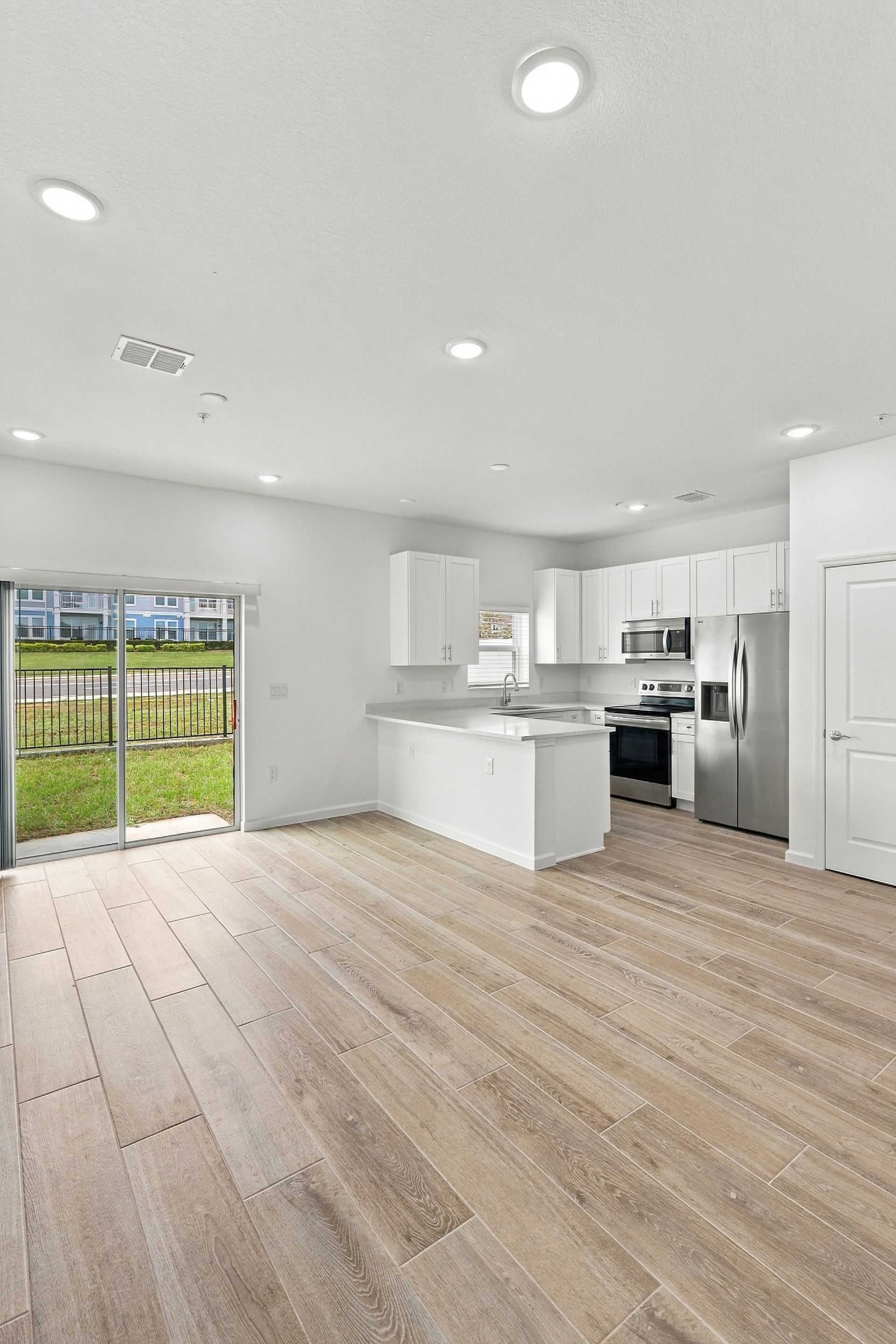 Bright, empty open-plan kitchen and living area with white cabinets, island, wood floors, and sliding glass doors