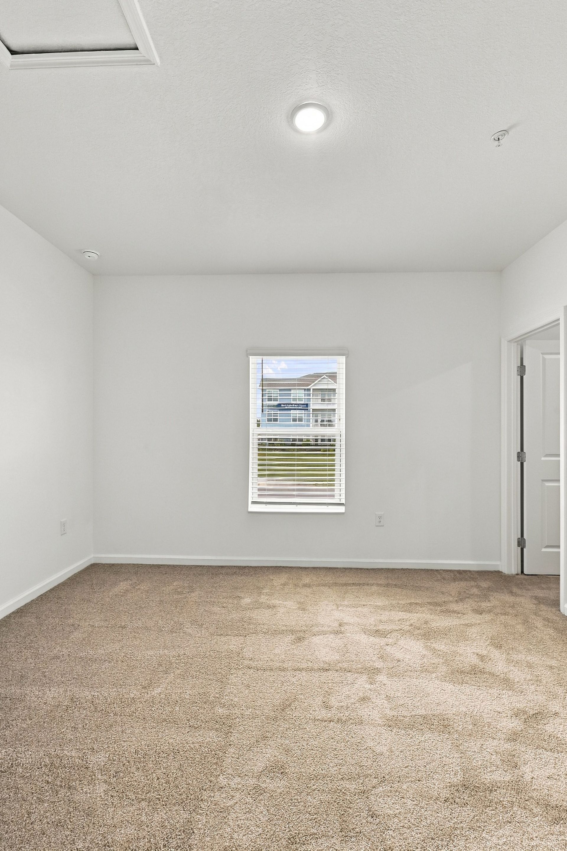 Empty white carpeted room with a window and ceiling light, viewed from the corner