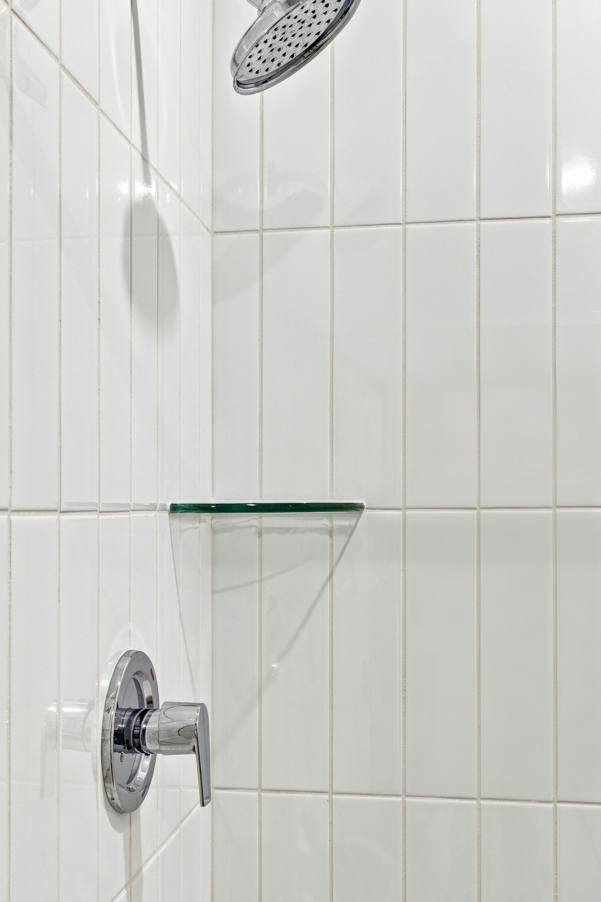 White tiled shower with chrome faucet and showerhead, plus a small glass corner shelf.
