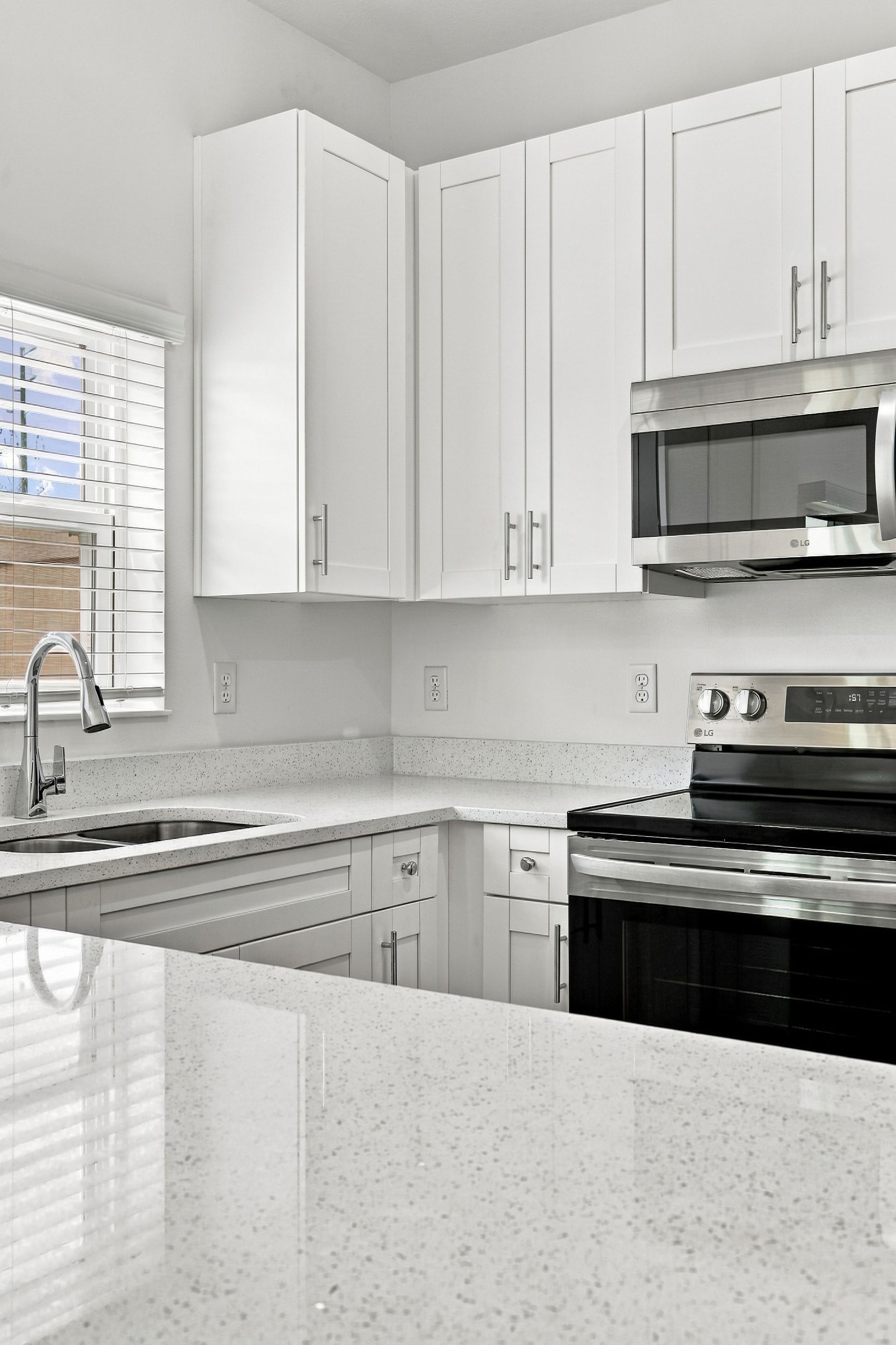 Bright white kitchen with stainless steel stove and microwave, corner sink, and speckled countertop