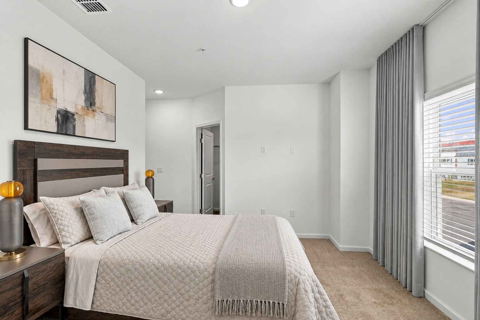 Bright bedroom with a bed, abstract wall art, white walls, and a large window with gray curtains.