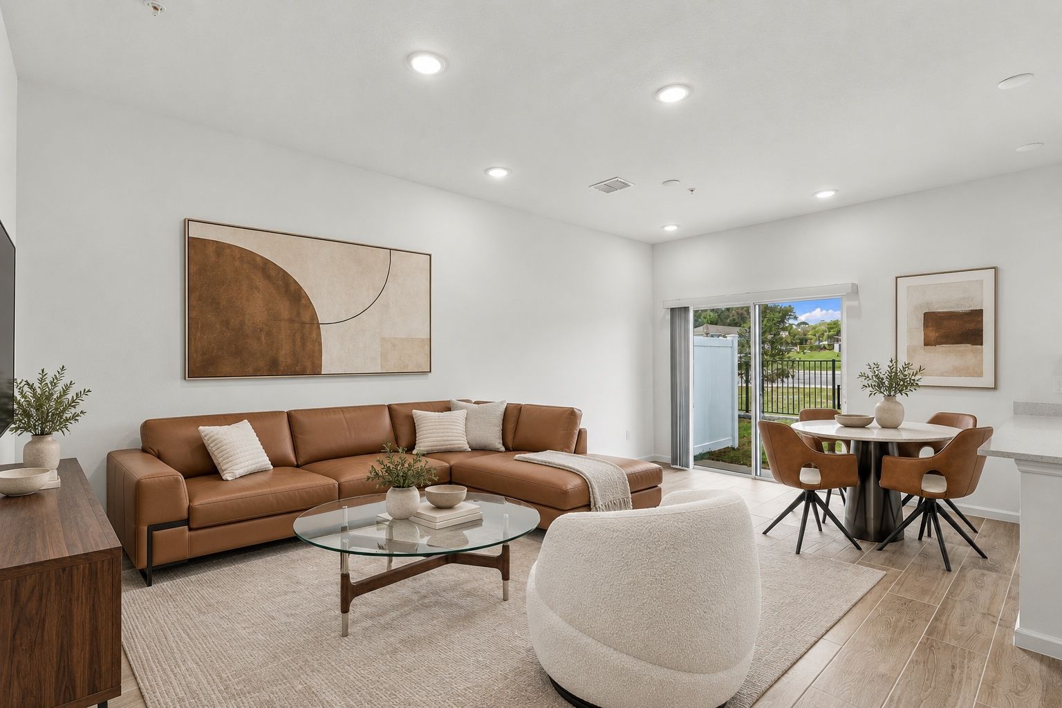 Bright modern living room with tan sectional, white chair, dining table, and large windows