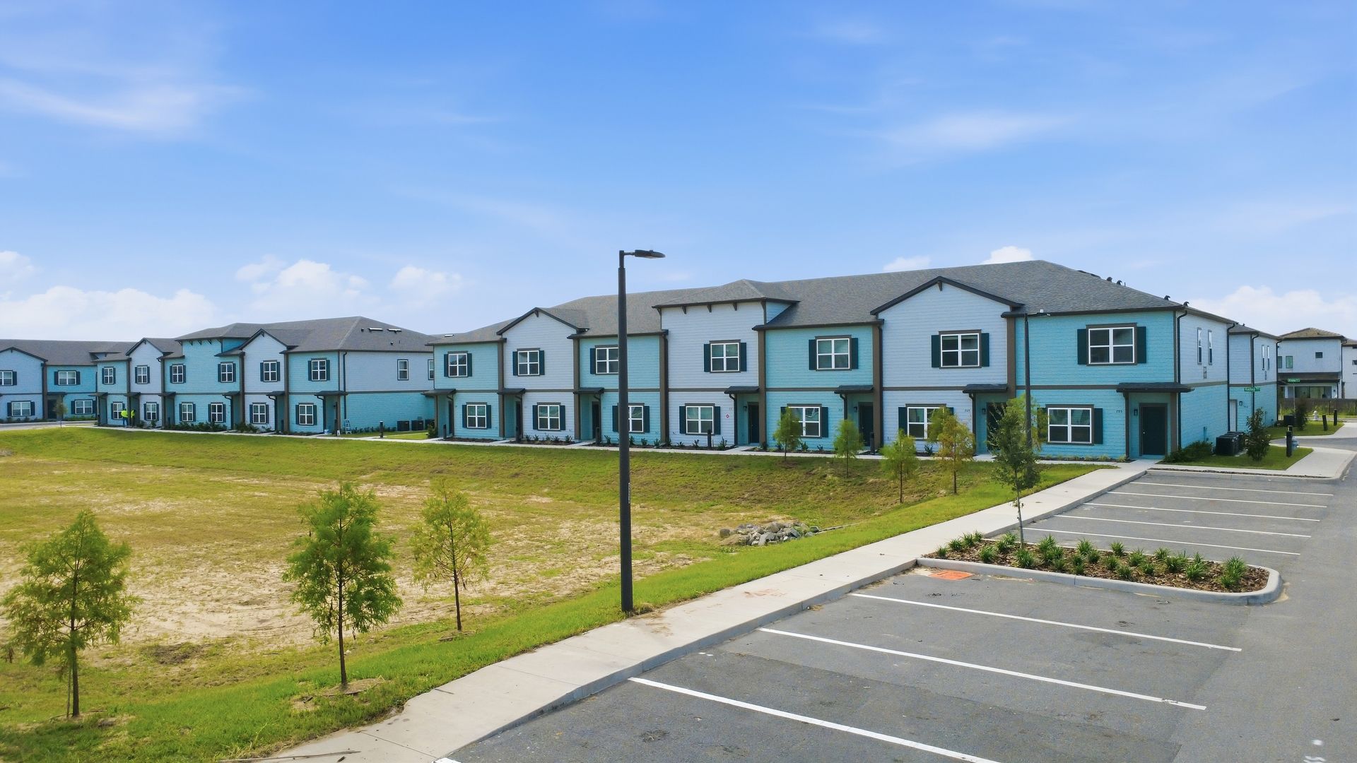 Modern apartment complex with blue siding beside a parking lot and grassy lawn under a clear sky