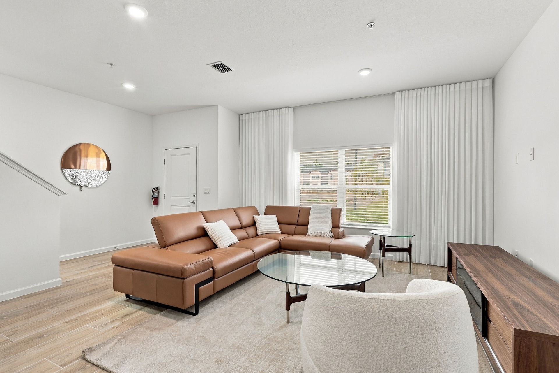 Bright modern living room with brown sectional, round coffee table, and large window with white curtains
