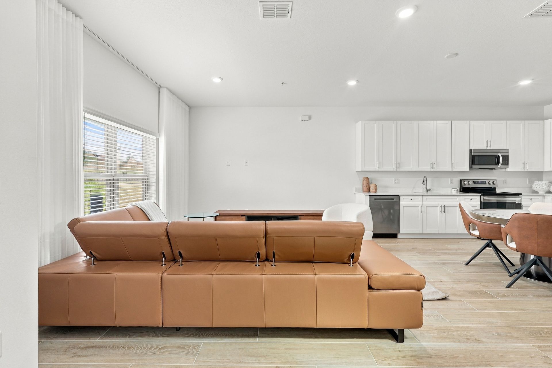 Bright open-plan living room with tan sofa, dining table, and white kitchen cabinetry