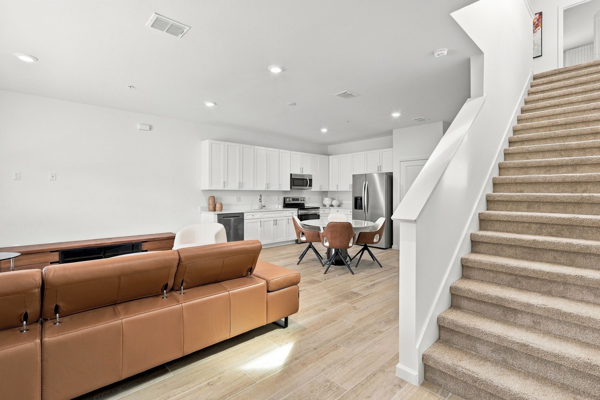 Open-concept living room with brown sectional, white kitchen, and carpeted staircase under bright lighting