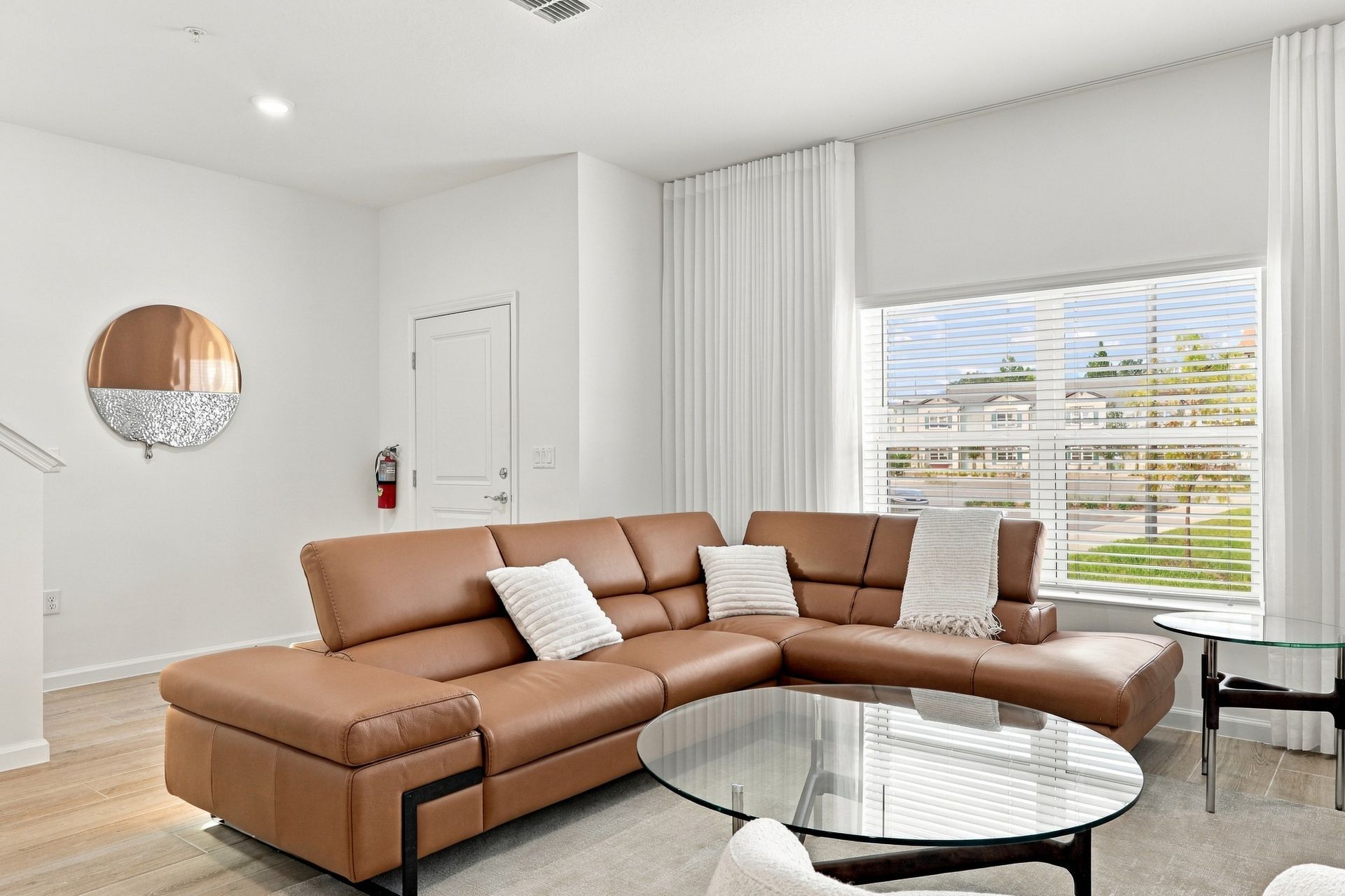 Modern living room with tan sectional sofa, glass coffee table, mirror, and large windows with blinds
