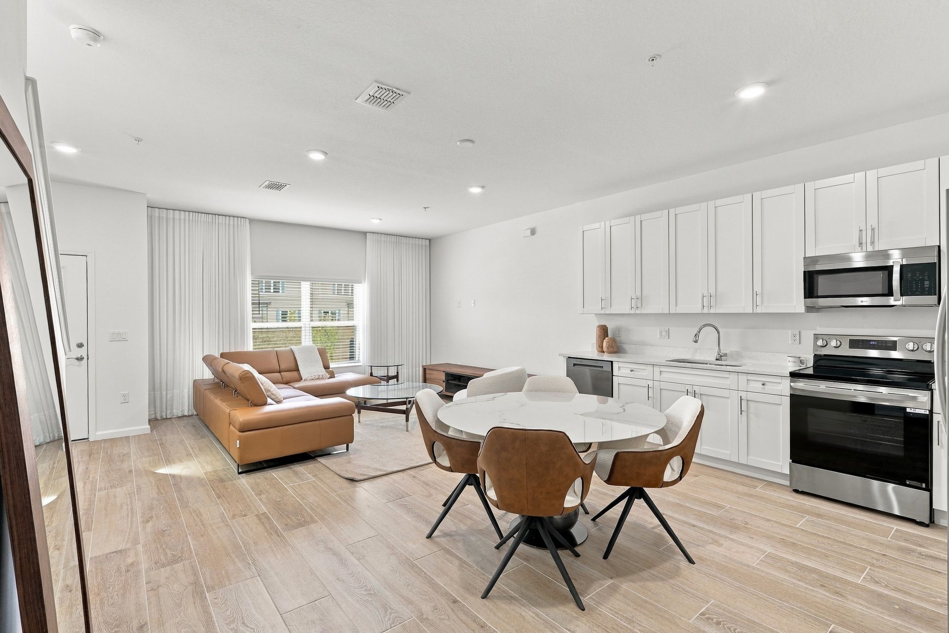 Bright open kitchen and dining area with white cabinets, wood floors, and tan sofa by large windows