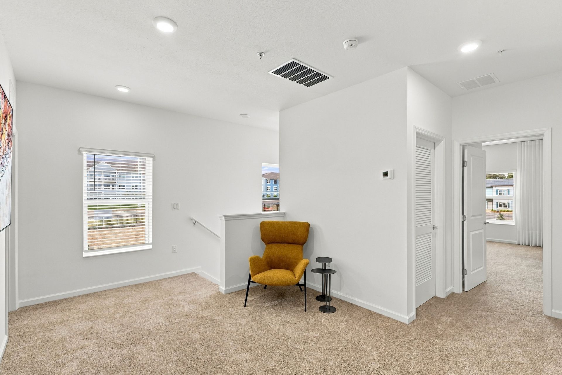 Empty beige-carpeted room with white walls, windows, a mustard chair, and a hallway opening to the right