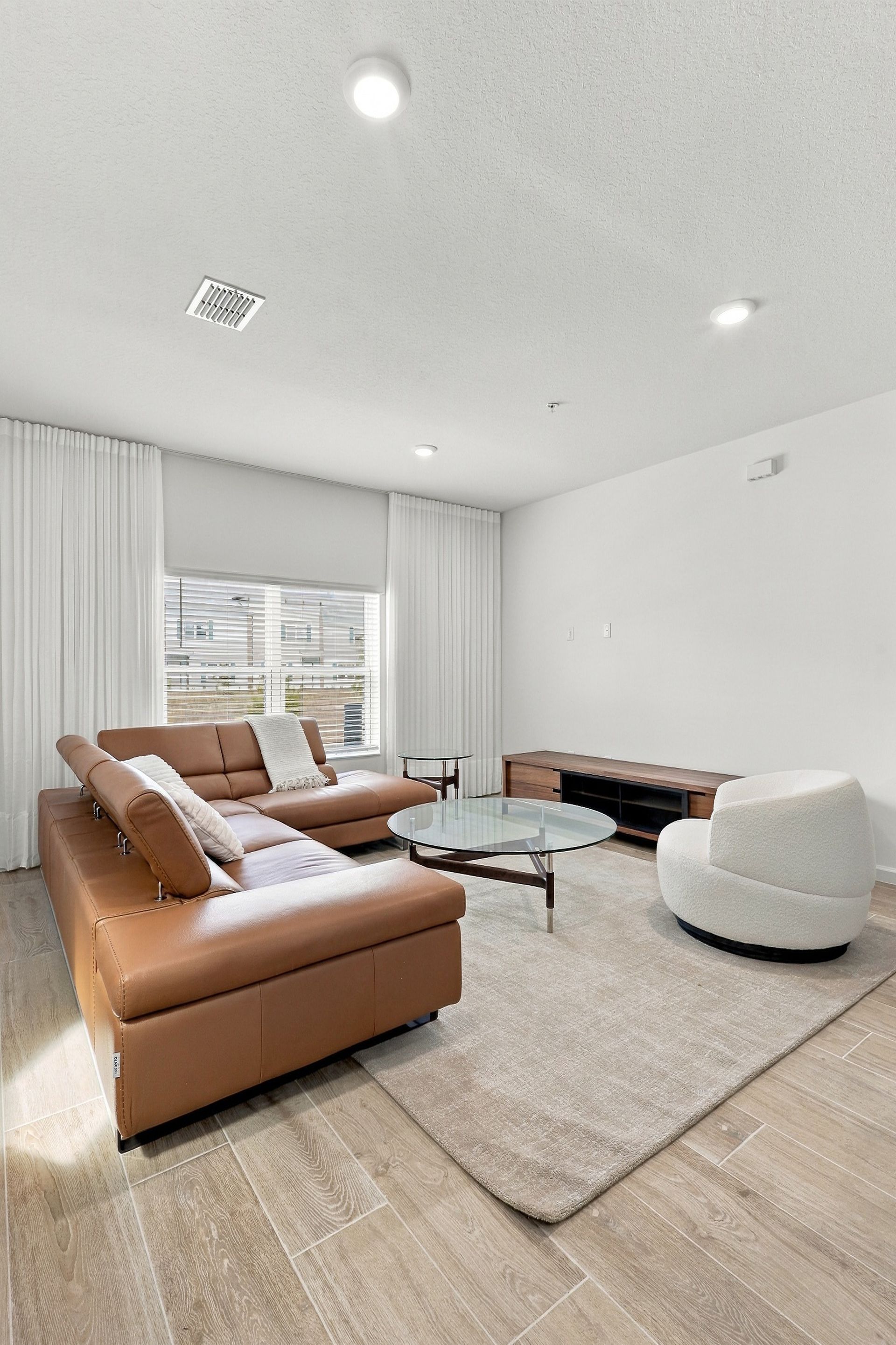 Bright living room with tan sectional, white chair, glass coffee table, and large window blinds
