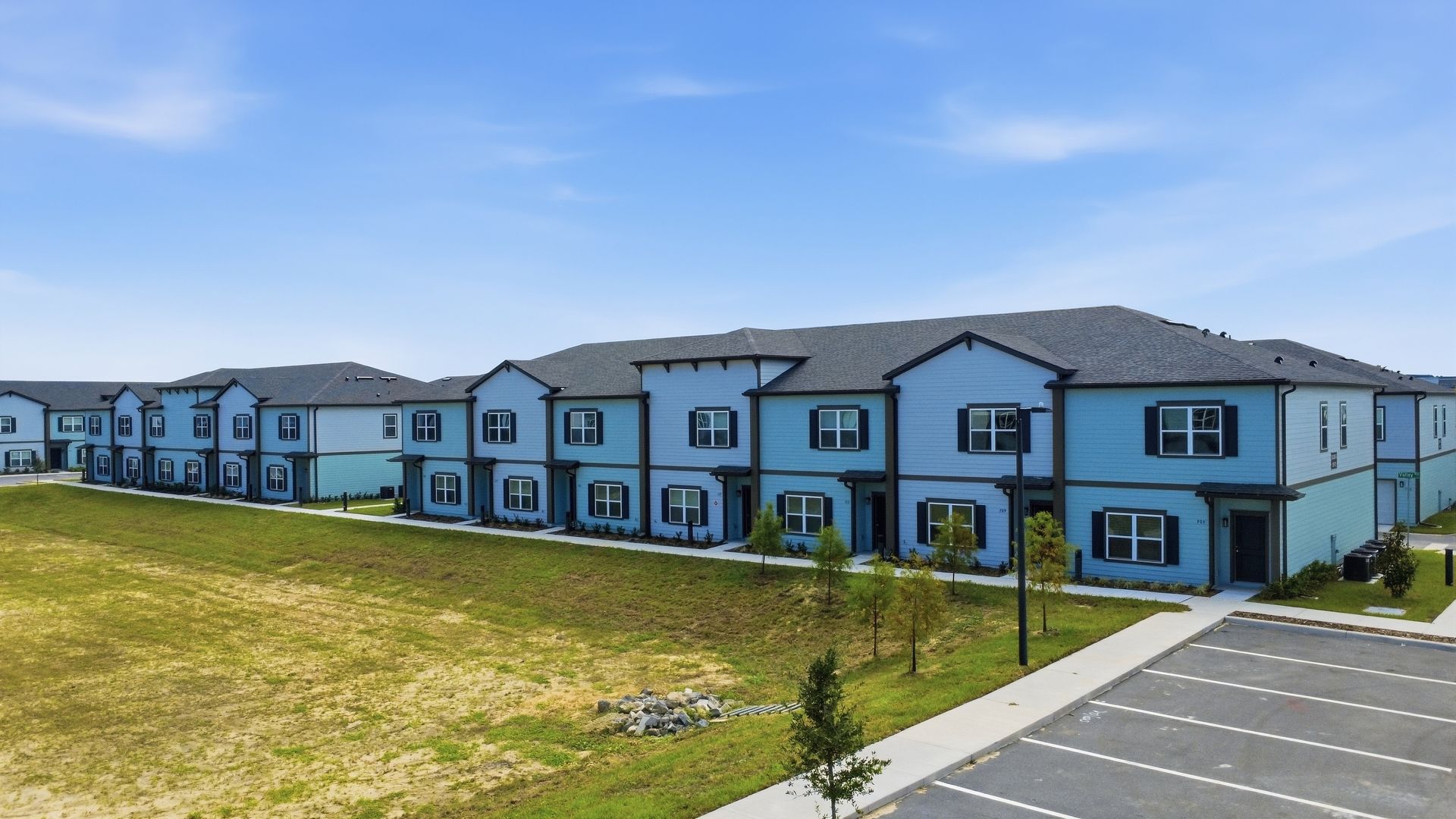 Row of light blue apartment buildings beside a grassy field and parking lot under a clear sky