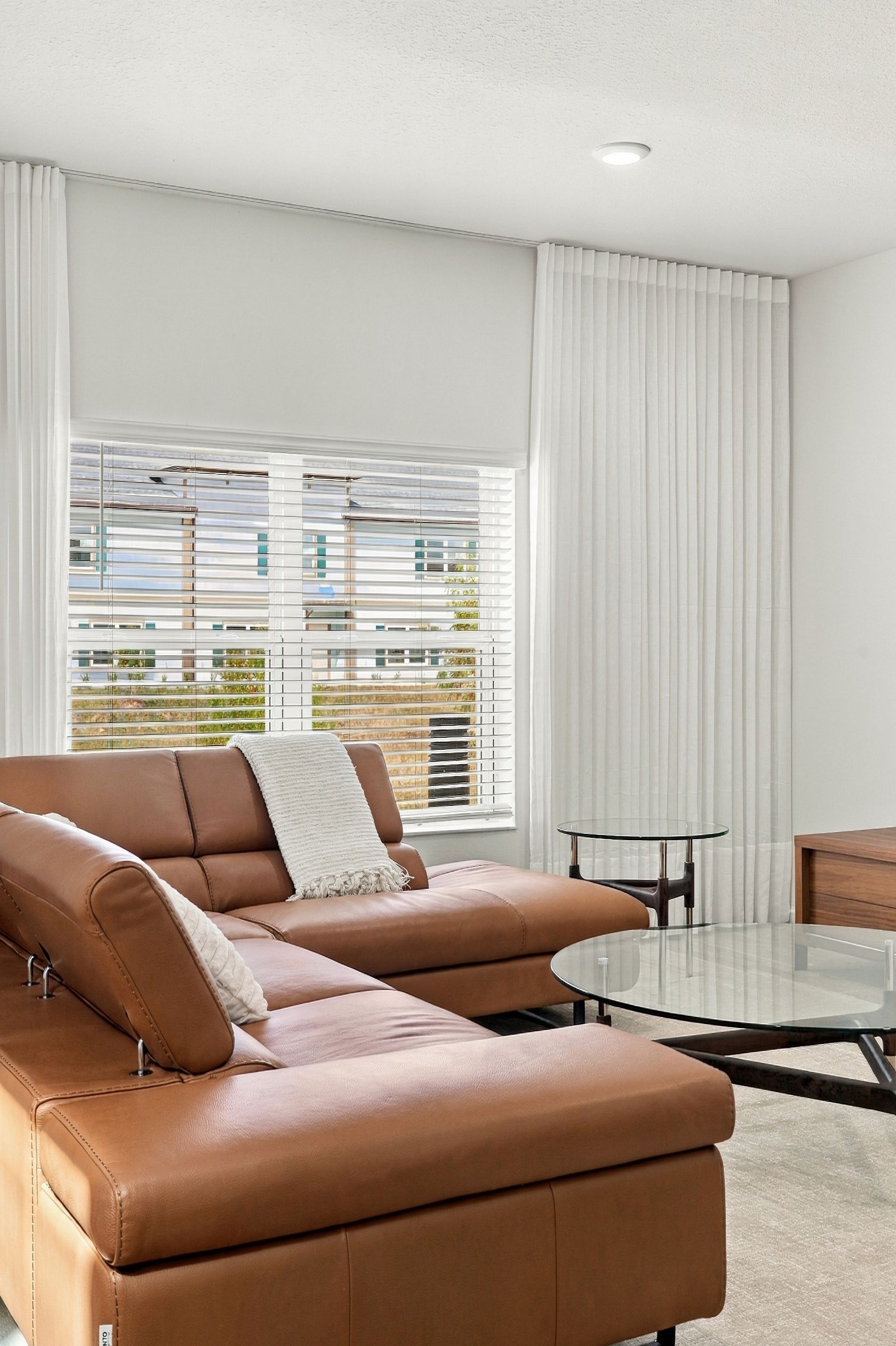 Bright living room with tan sectional, glass coffee table, white curtains, and large window.