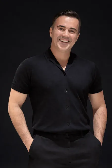 Smiling person in a black short-sleeve shirt and pants, standing with hands in pockets against a dark background