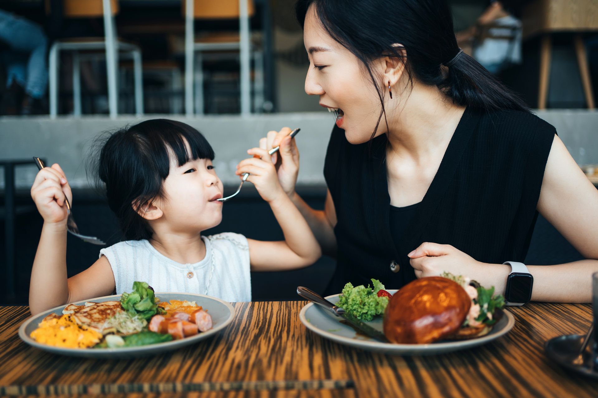 Woman feeding child at a table in a restaurant; both eating from plates of food with forks.