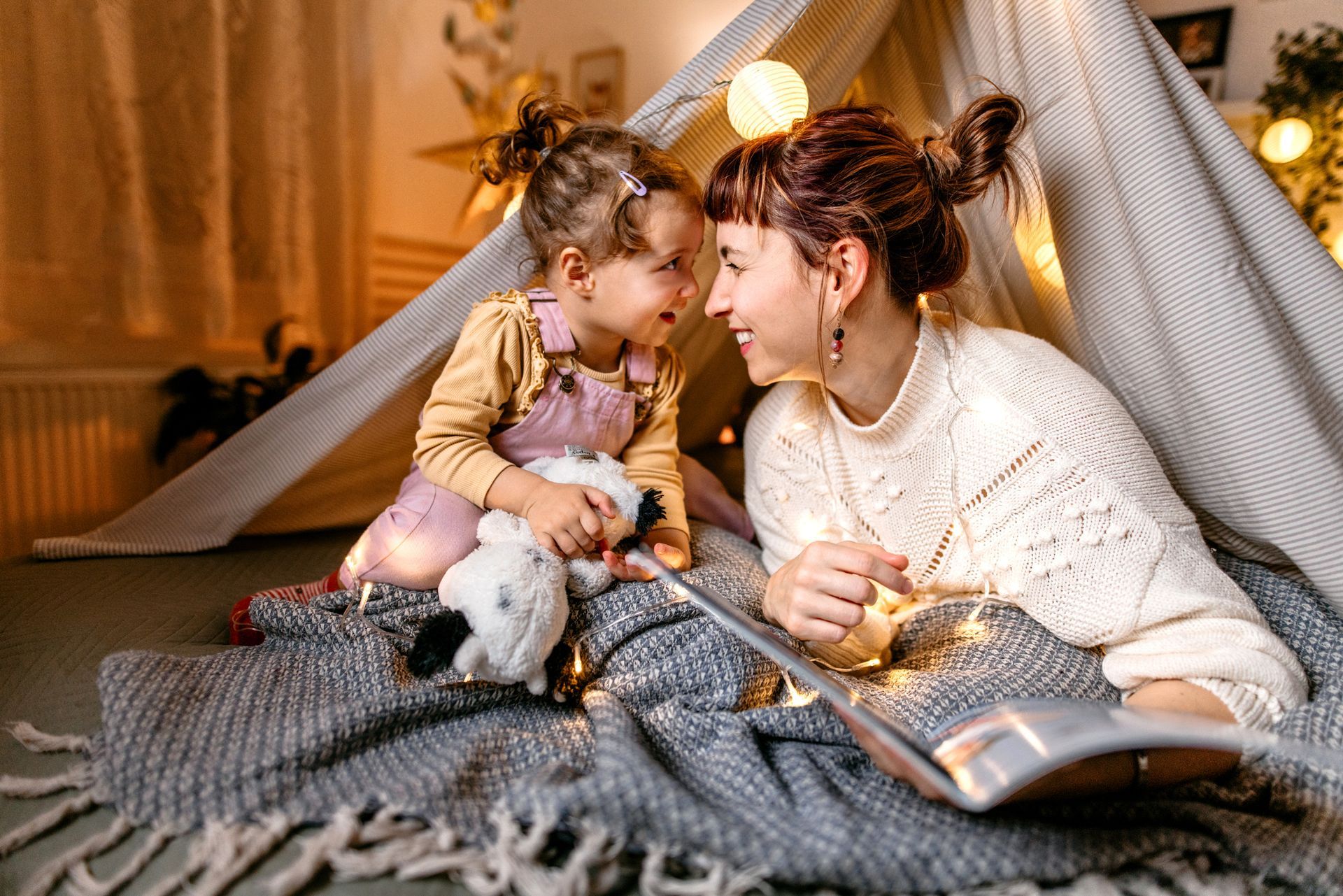 A woman and a little girl are sitting under a tent reading a book.