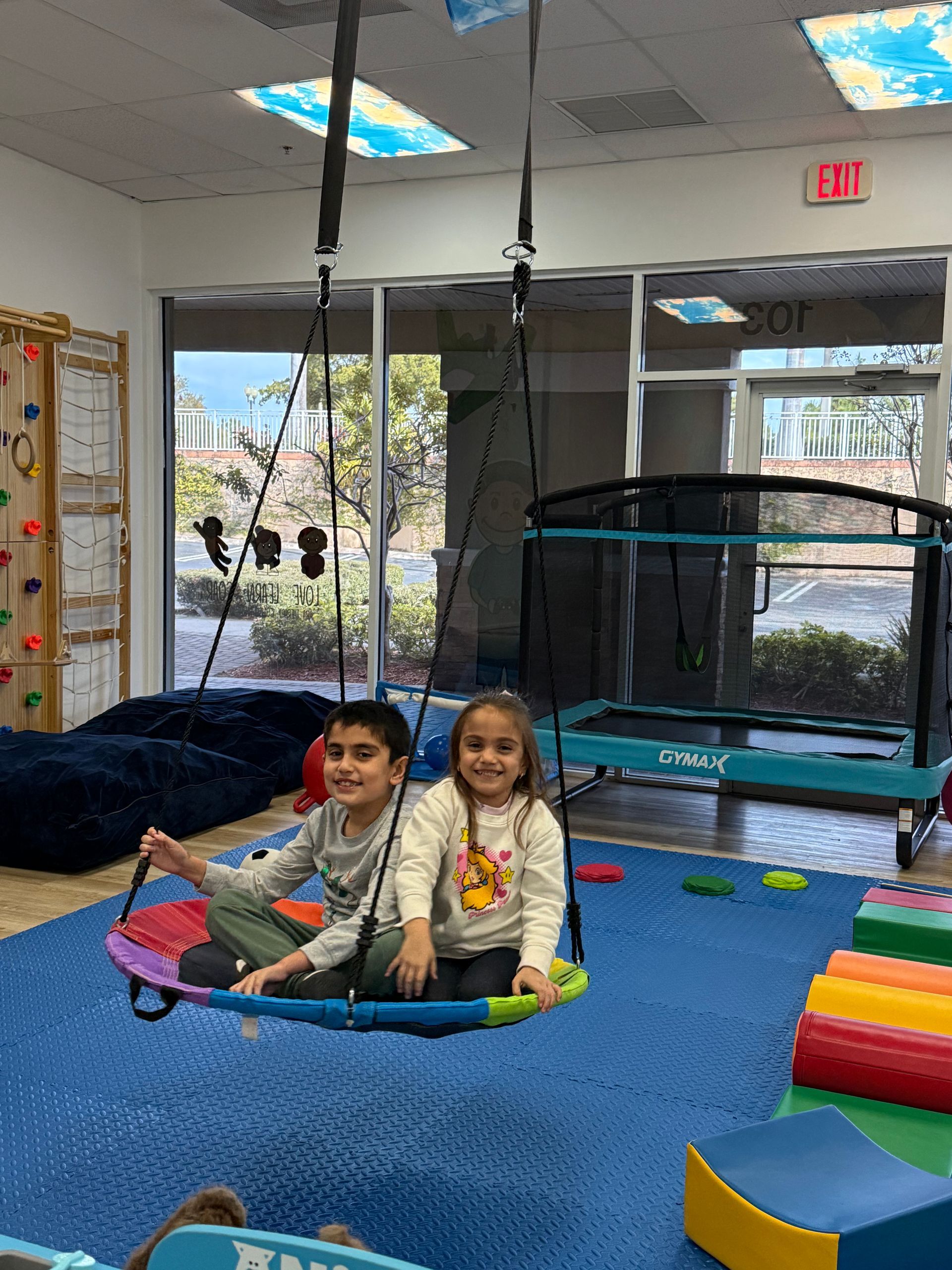 Two children sit on a swing in a play area. They smile. There’s a trampoline and climbing wall nearby.