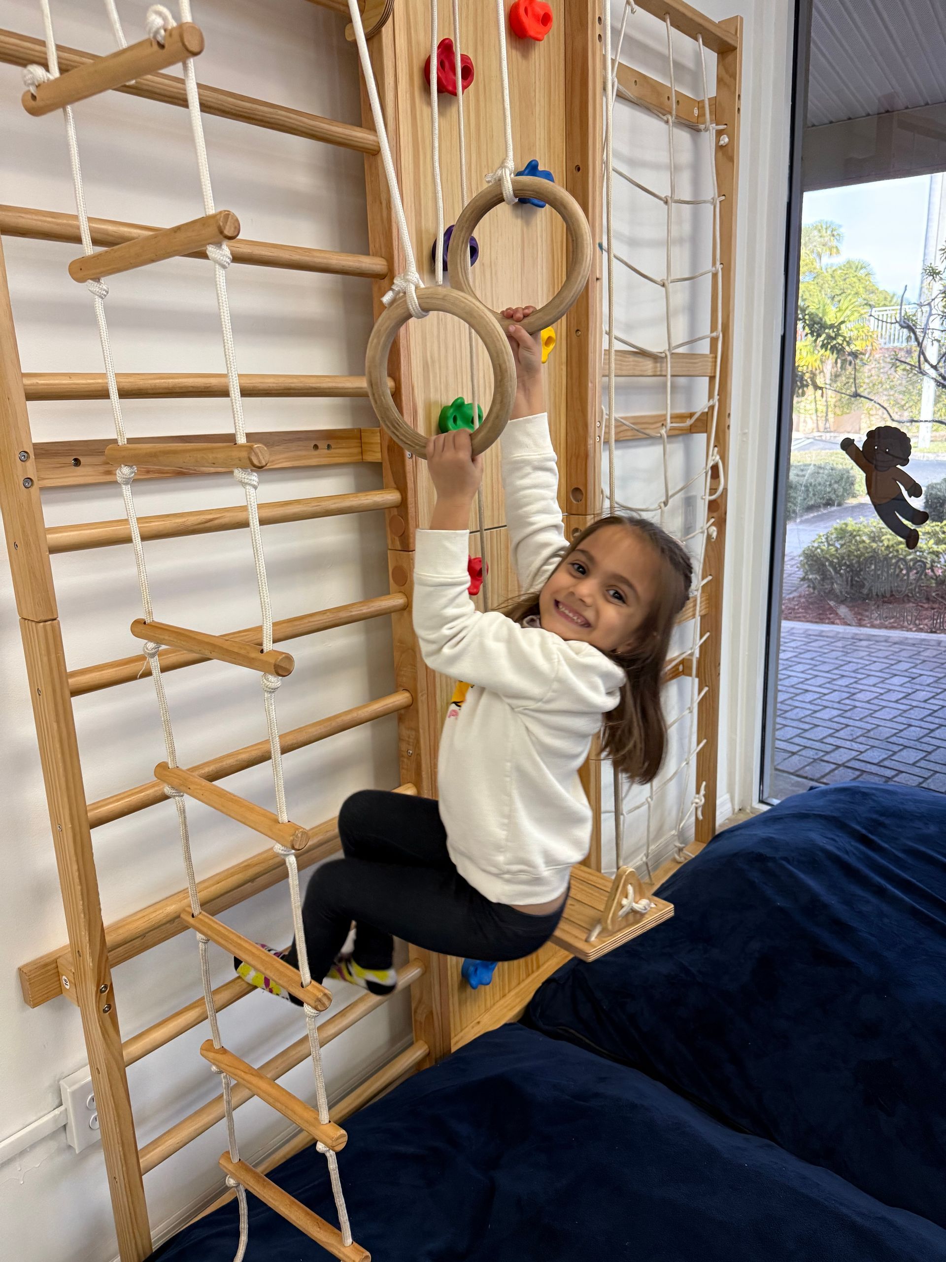 Girl on wooden climbing structure with rings and ladder, smiling, indoor.