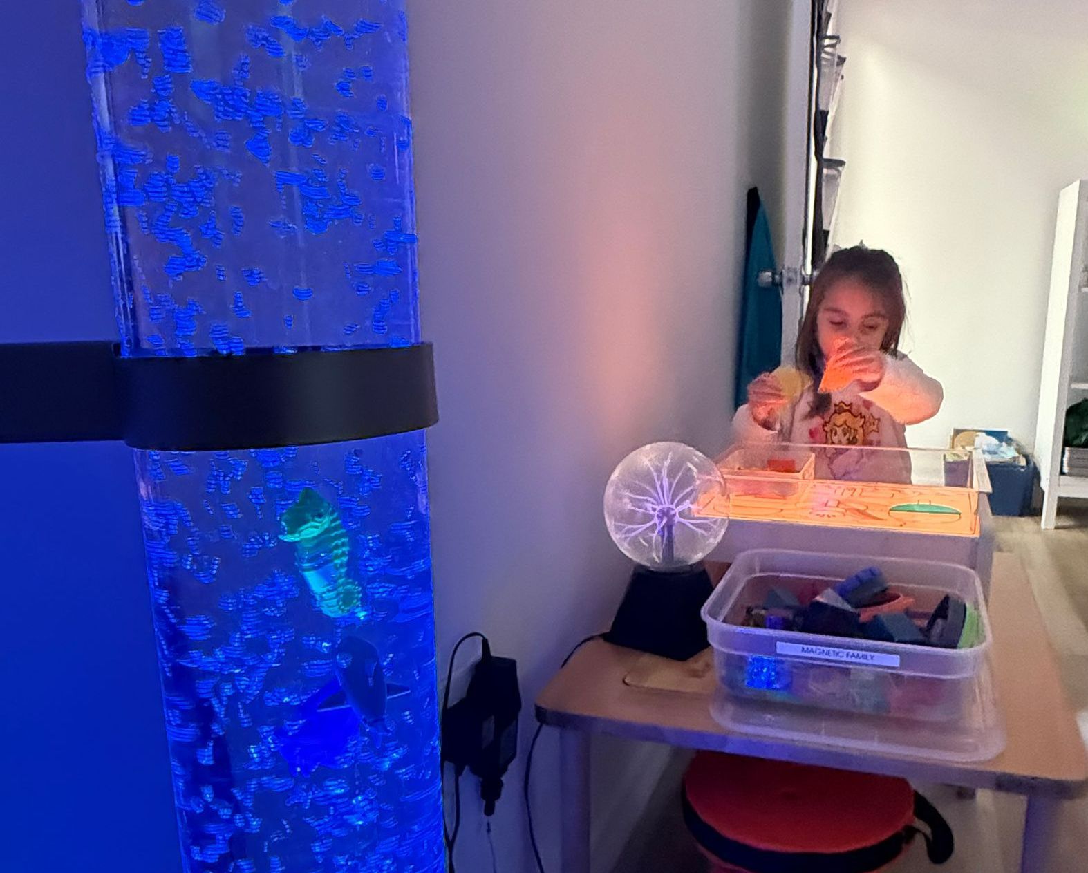 Child interacting with sensory items, including a bubble tube and a plasma ball, at a table.