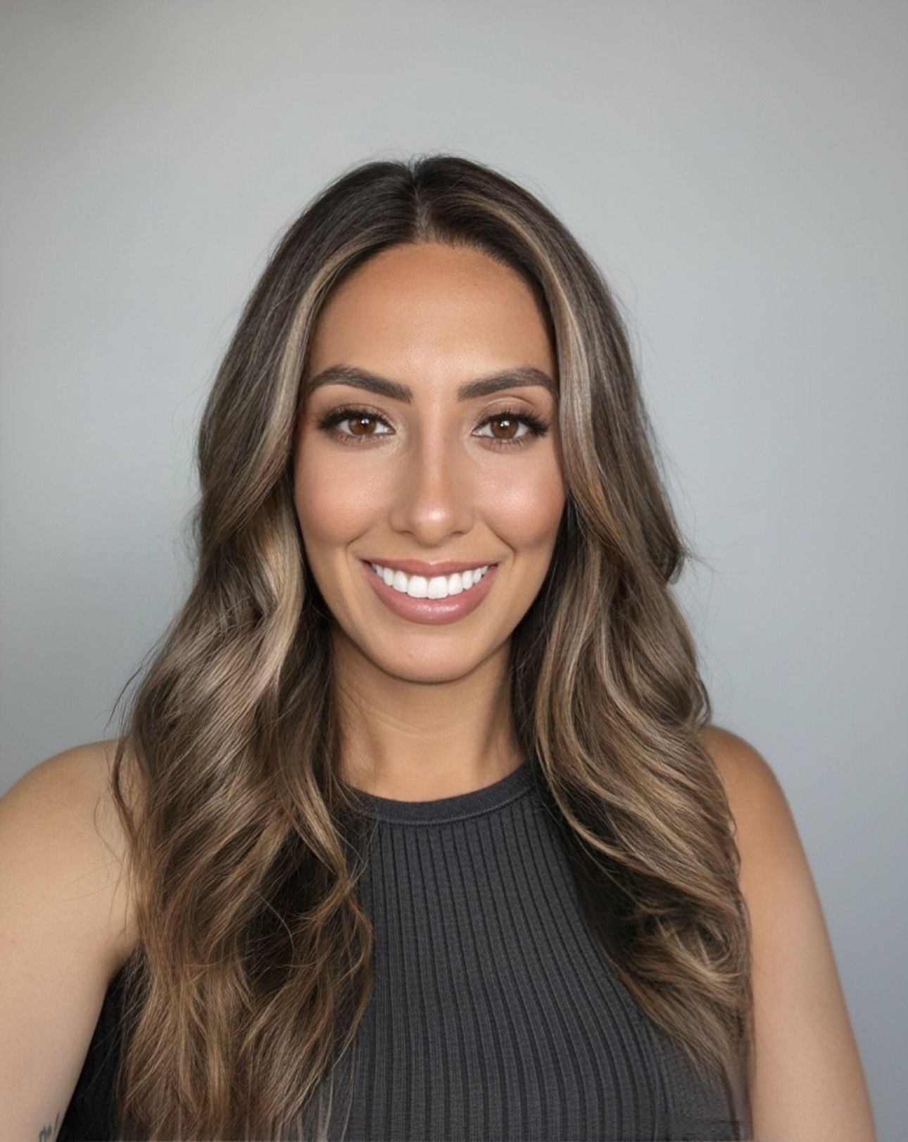 Smiling woman with long wavy brown hair wearing a dark sleeveless top against a gray background