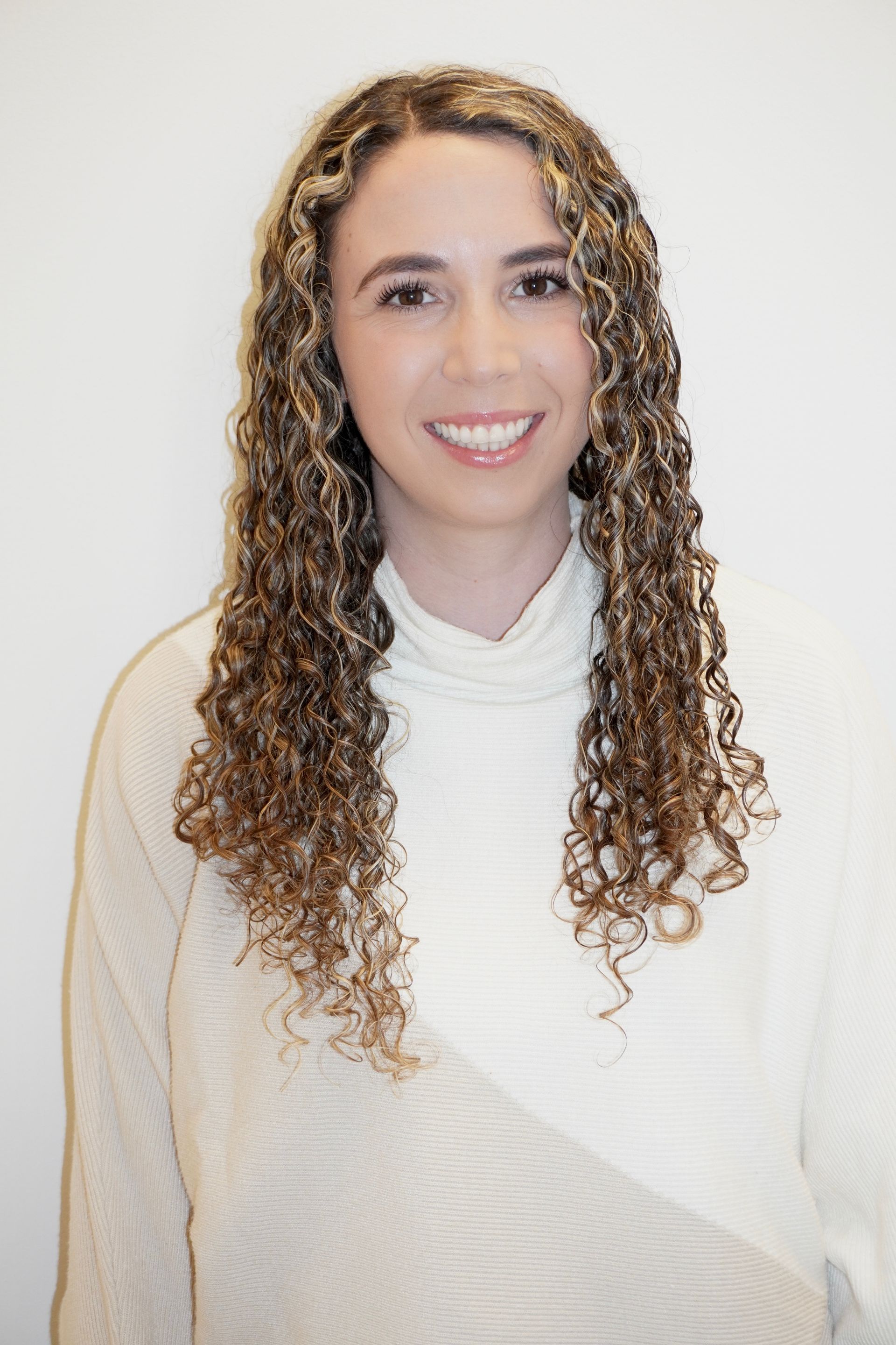 Woman with curly brown hair, wearing a white turtleneck sweater, smiling against a white background.