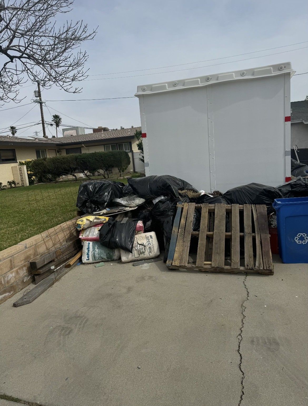 Pile of trash bags, cardboard boxes, and a wooden pallet by a large white structure and blue recycling bin on a driveway.