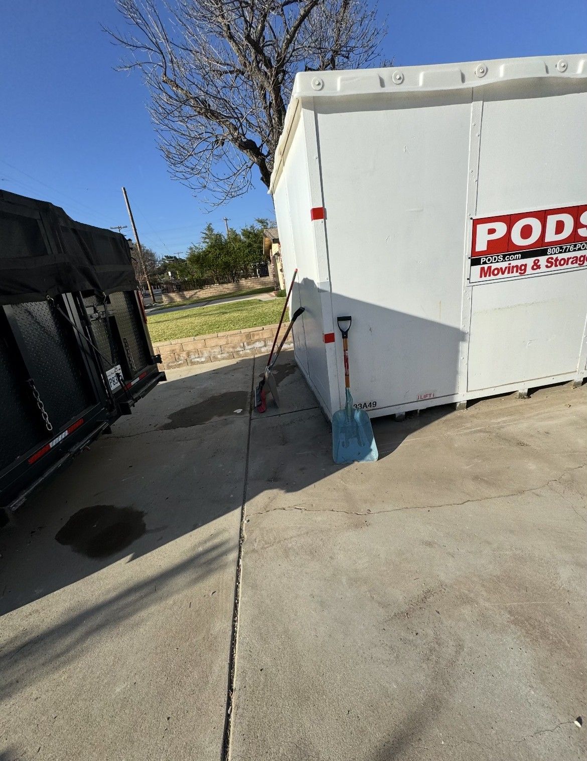 PODS storage container next to a black trailer on a concrete surface, with tools and a tree in the background.