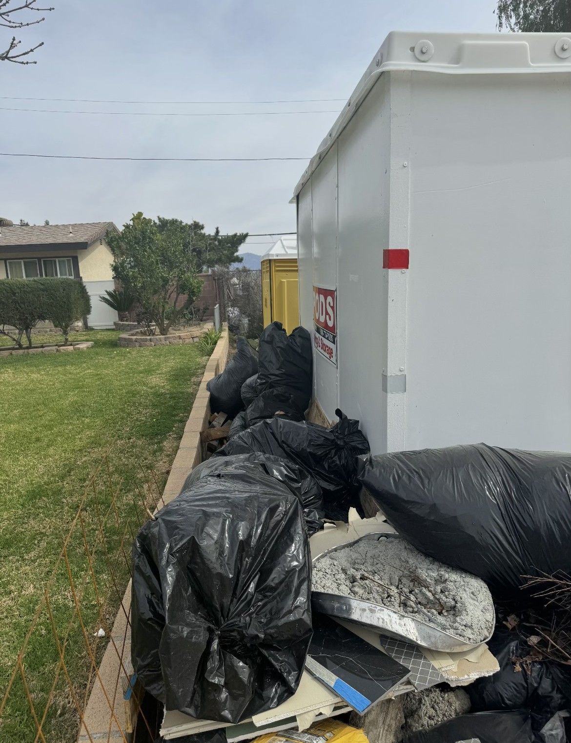 Black trash bags and debris piled next to a large white dumpster in a residential area.