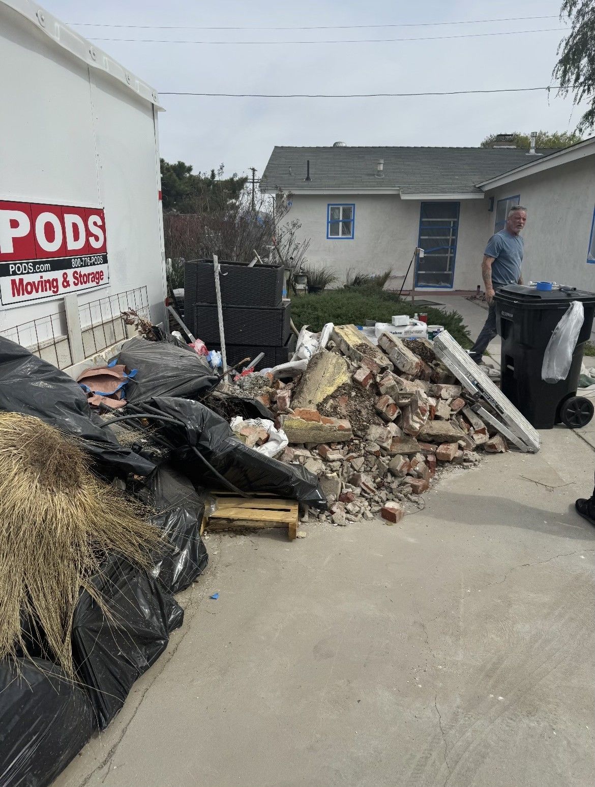 Pile of construction debris and trash on a driveway, next to a PODS container and a house; a person walks by.